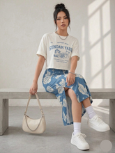 Load image into Gallery viewer, A woman sitting on a bench wearing a t-shirt, a blue floral skirt, and white sneakers.