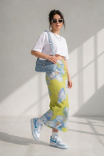 Load image into Gallery viewer, Woman wearing a white crop top, yellow floral pants, and blue sneakers in a sunlit room.