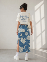 Load image into Gallery viewer, Person wearing a white t-shirt with text and graphics, and a blue floral skirt in a bright room.
