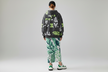 Load image into Gallery viewer, Person wearing a black hoodie with colorful designs, green leaf-patterned pants, and white sneakers on a gray background
