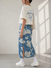 Load image into Gallery viewer, Person wearing a white t-shirt with text and a blue floral skirt in a bright room.
